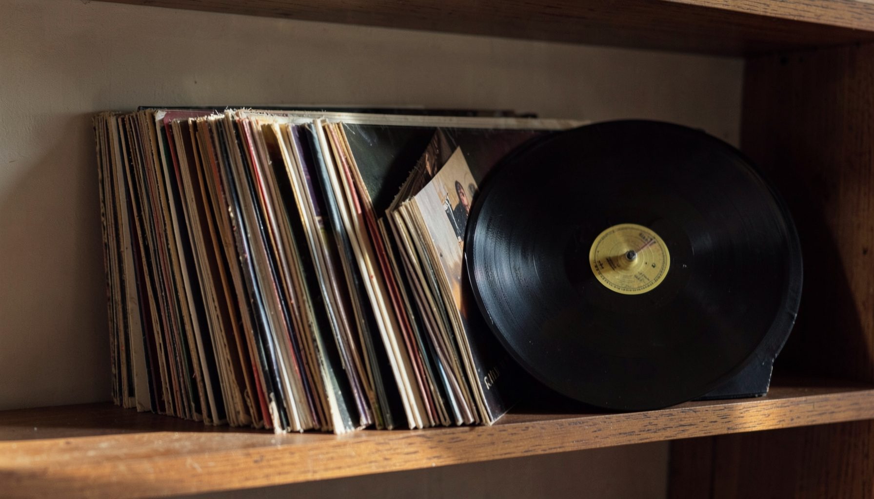 Collection of vinyl records on a wooden shelf, with one record removed from its sleeve, dark warm tones, nostalgic mood