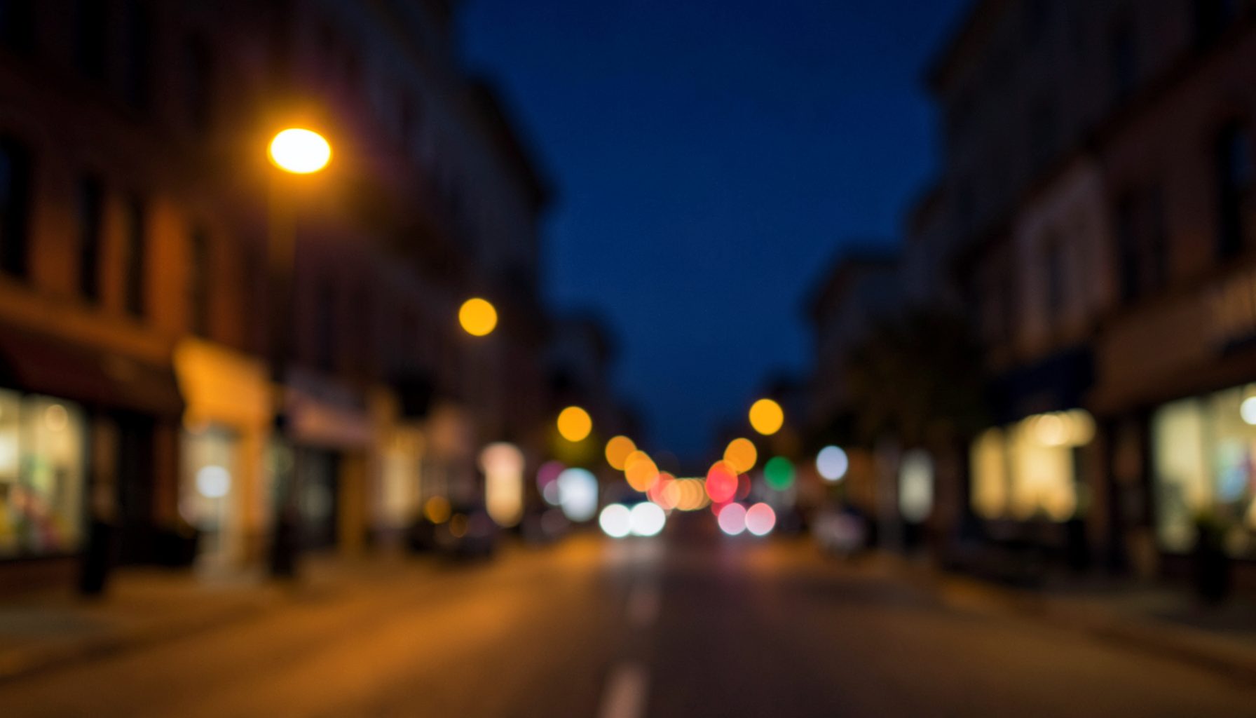 Blurred night cityscape of a street lined with buildings and glowing lights, warm and cool tones, quiet mood